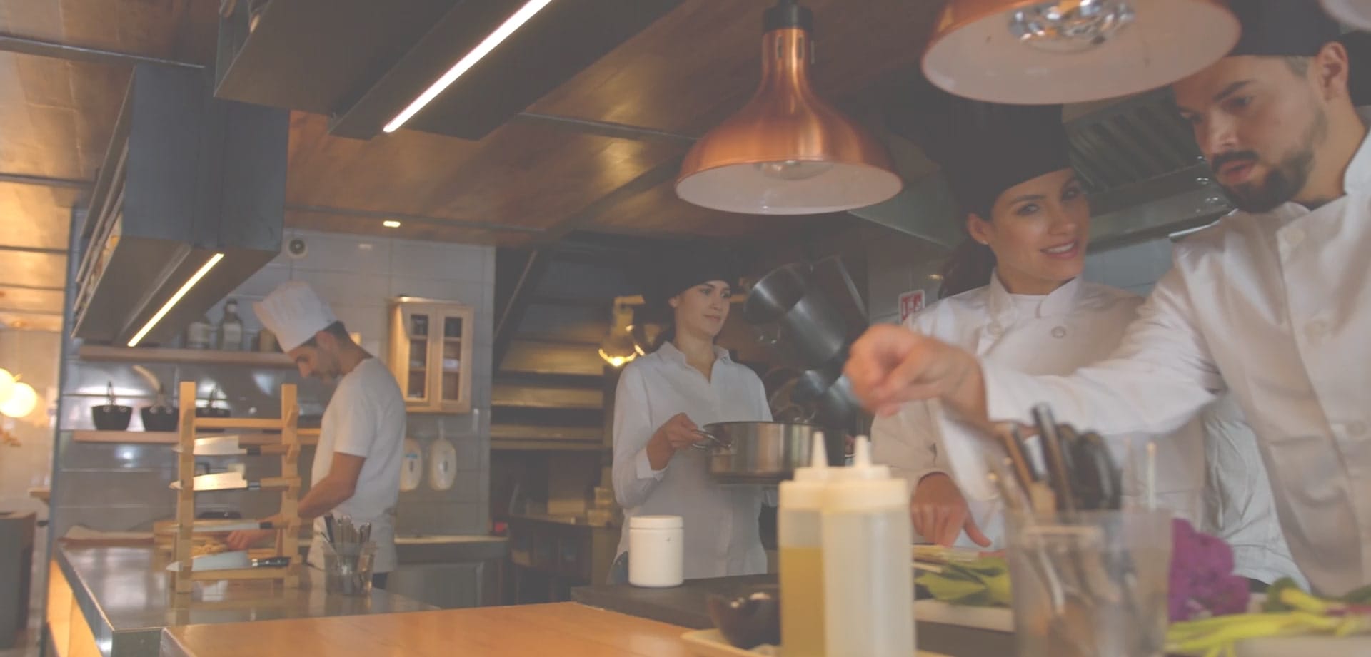 culinary-academy-of-las-vegas-background-1 Chefs working together in a commercial kitchen, preparing food under hanging copper lights