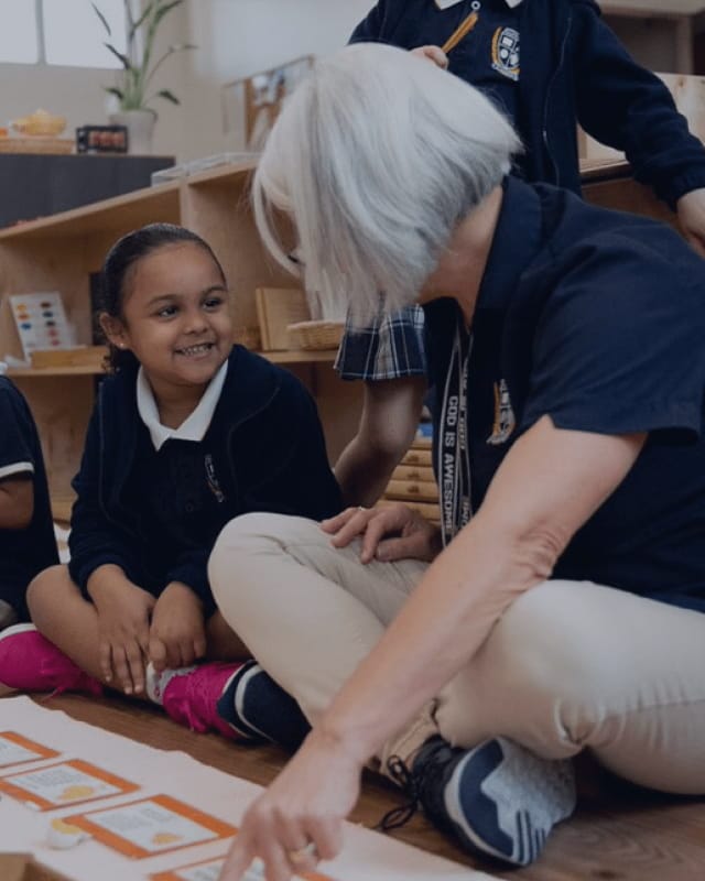 A teacher sitting on a classroom floor working through a learning activity with a smiling young student in school uniform.