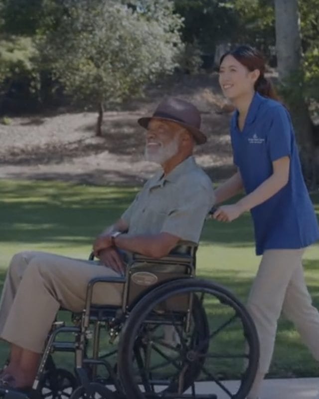 digital-marketing-agency-healthcare Caregiver pushing an elderly man in a wheelchair along a paved path in a sunny park.